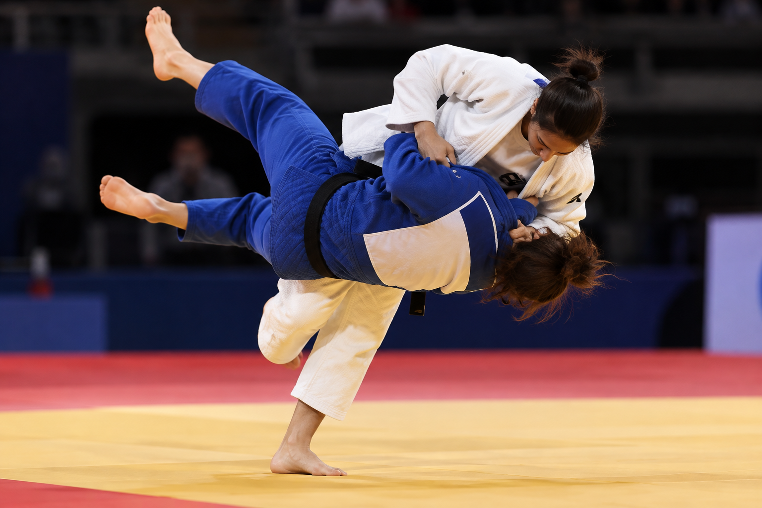Duas judocas em combate durante uma luta de judô. A atleta de quimono branco aplica um golpe de uchi mata e ergue a adversária de quimono azul no ar, em um momento de ação congelado sobre o tatame ama